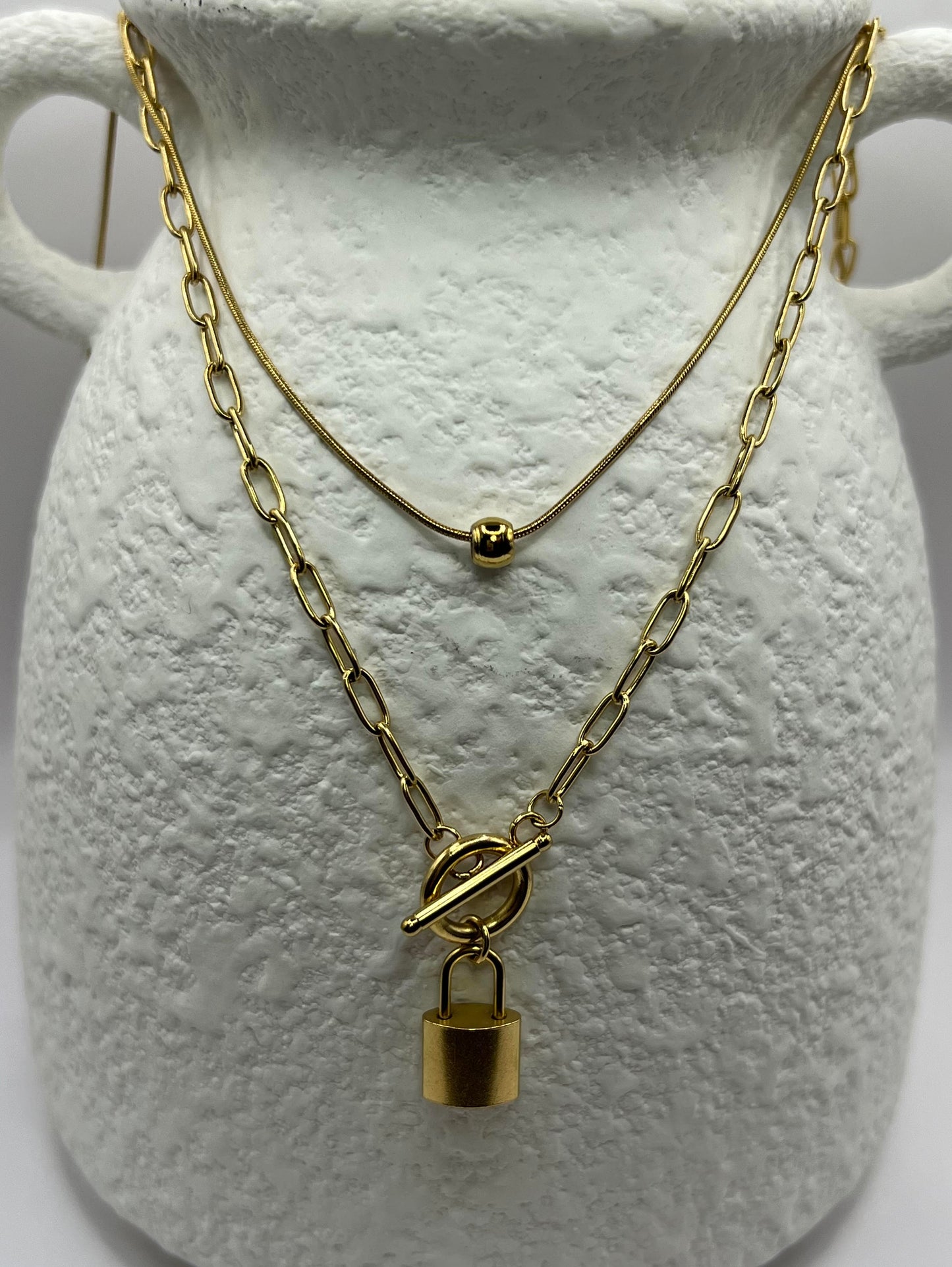 Two gold necklaces with a lock pendant on a textured white surface