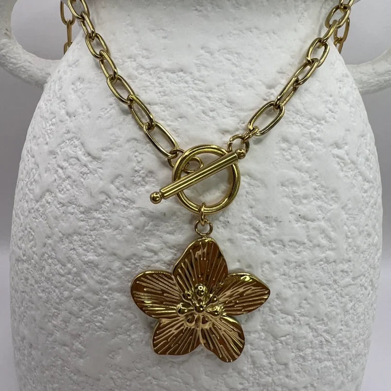 A gold-colored necklace with a floral pendant, displayed on a white stand.
