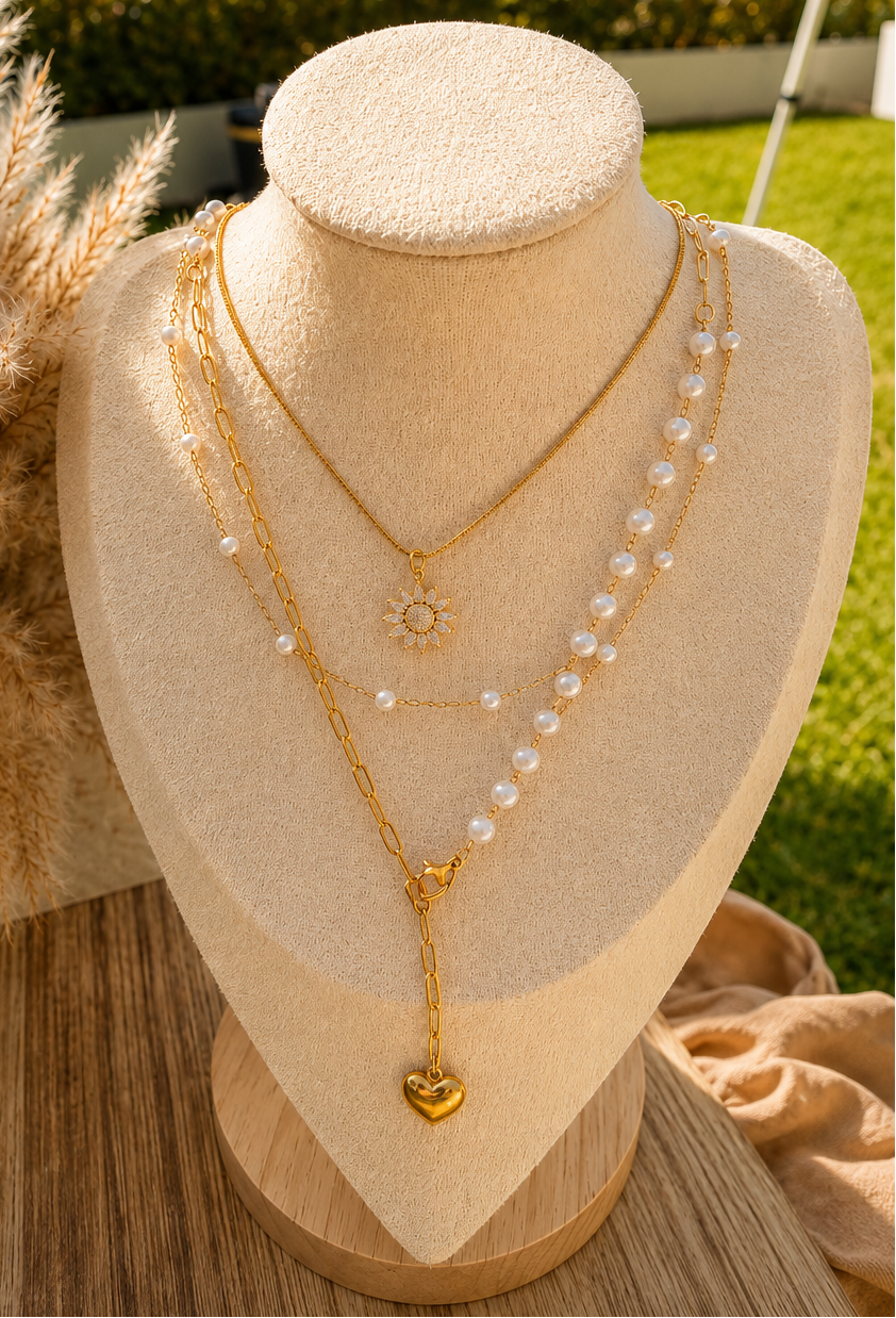 A layered gold necklace set displayed on a beige jewelry bust outdoors. The set includes a delicate gold chain with a small sunburst pendant, a longer gold chain with a heart-shaped charm, and a strand of white faux pearls.