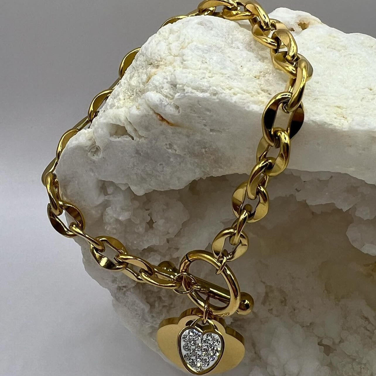 A gold-colored bracelet with a heart-shaped charm, displayed on a rock crystal.