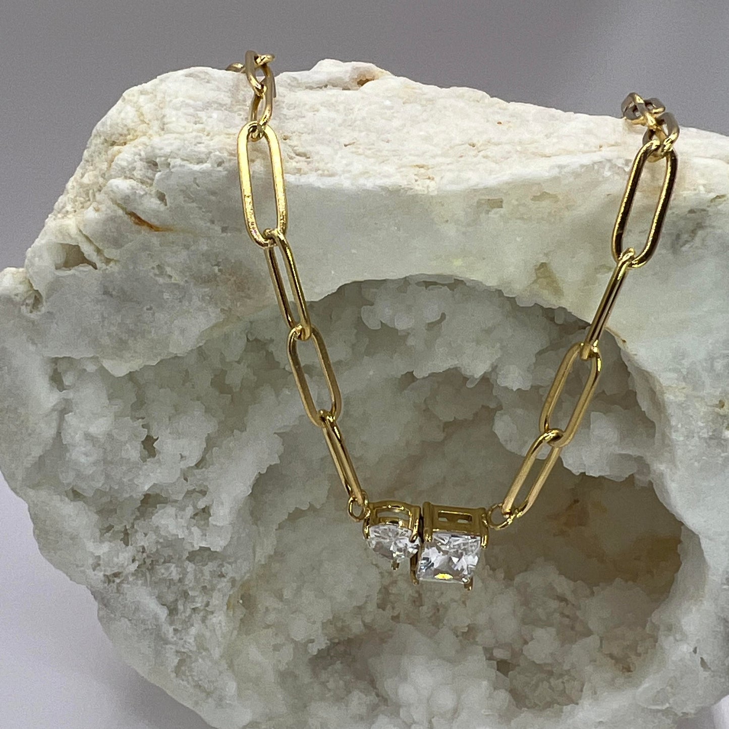 A gold-colored necklace with a pendant featuring a cubic zirconia surrounded by smaller clear crystals, displayed on a rock.