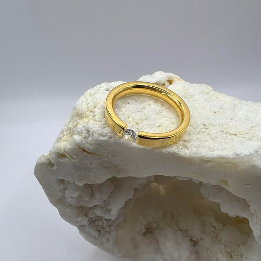 A gold ring with a clear, square-cut gemstone on top, placed on a textured white rock.